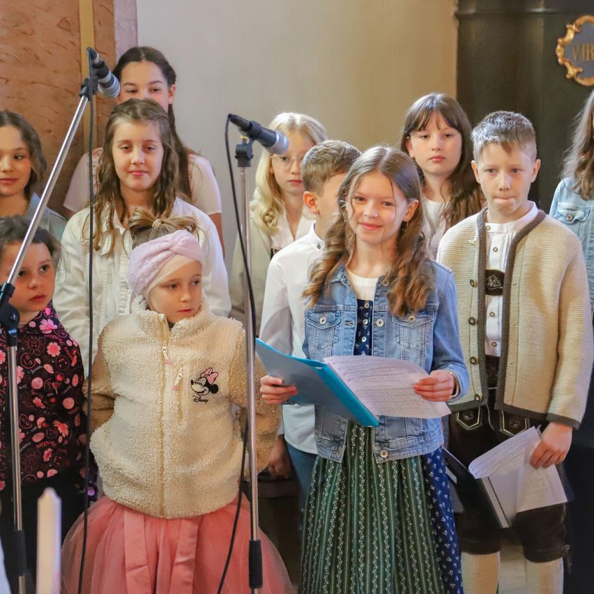 A choir of children, some holding music sheets, sing into a microphone in a church. A girl with a headband looks at the camera.