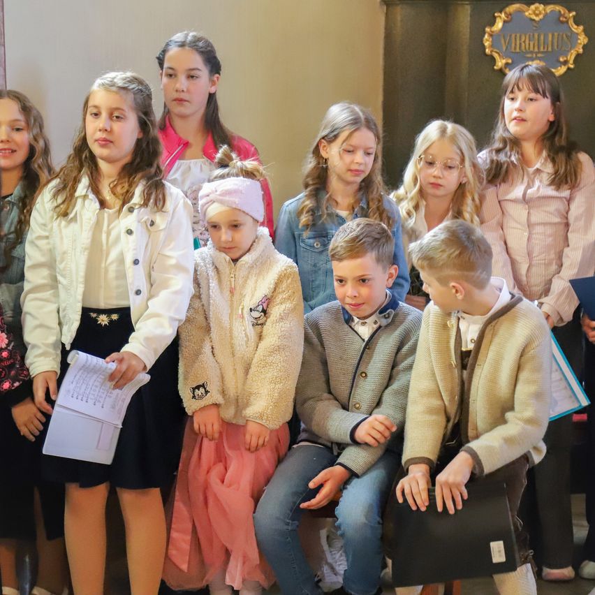 A group of children, boys and girls, are standing in a line, possibly for a photo. They are dressed in various outfits, some with glasses, and holding papers. The children in the front row are looking at something. Behind them is a wall with an emblem.
