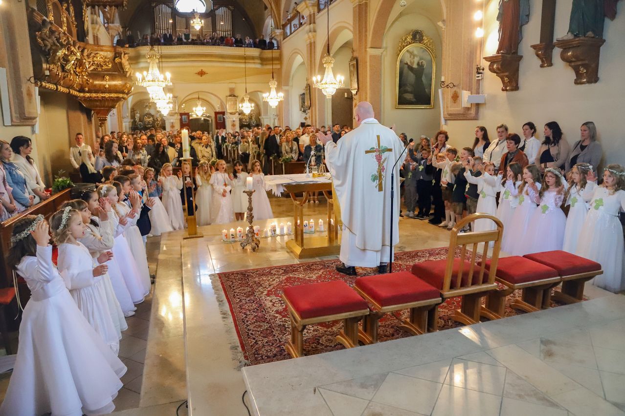 A priest delivers a sermon at a church. Children in white dresses stand nearby, and a crowd is gathered. Candles, a microphone, and a cross adorn the altar.
