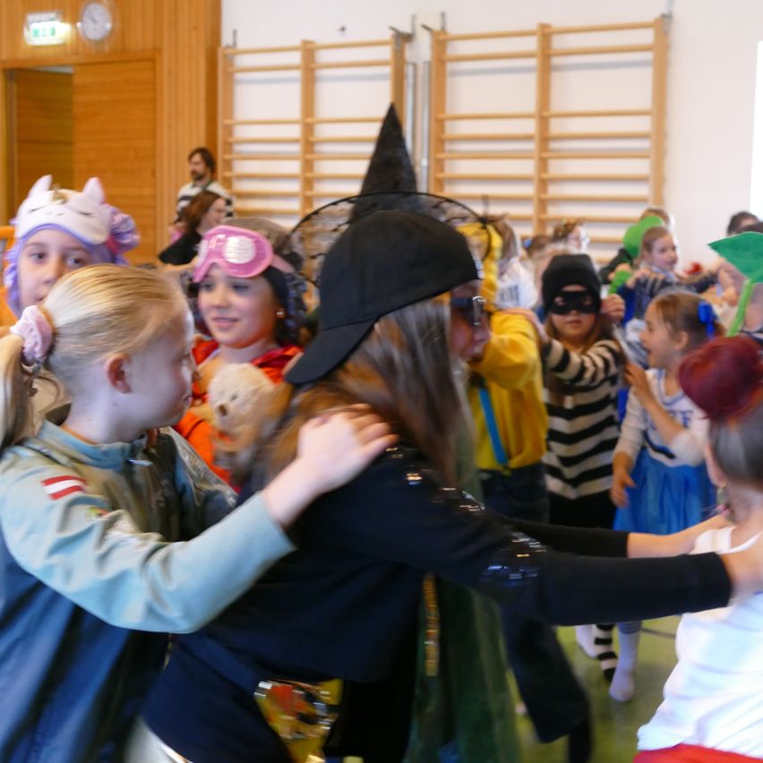 Eine Gruppe von Kindern in Kostümen ist drinnen versammelt, einige tragen Masken. Ein Mädchen mit einer Hexenmütze umarmt ein anderes Kind.