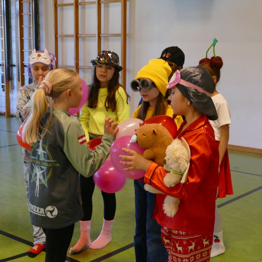 Eine Gruppe von Kindern ist in einem Raum versammelt, einige in Kostümen. Ein Mädchen hält Luftballons und einen Teddybären, während andere Hüte und Brillen tragen. Der Raum hat weiße Wände und grünen Boden.