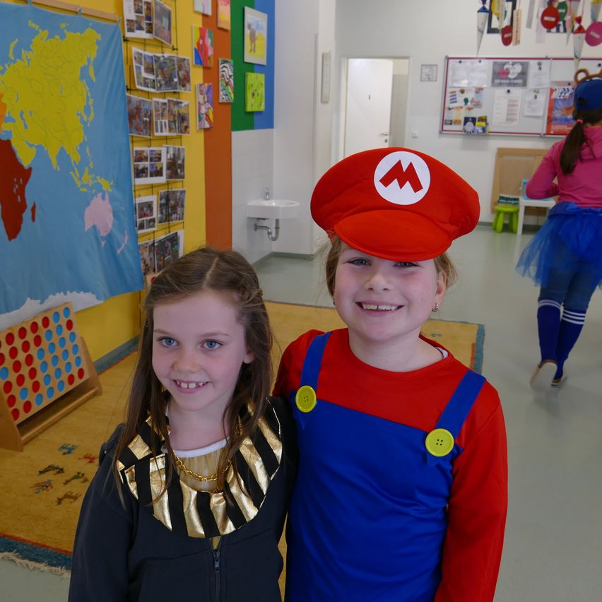 Zwei junge Mädchen, eine als Mario und die andere als ägyptische Königin verkleidet, stehen in einem Klassenzimmer mit einer Weltkarte an der Wand.