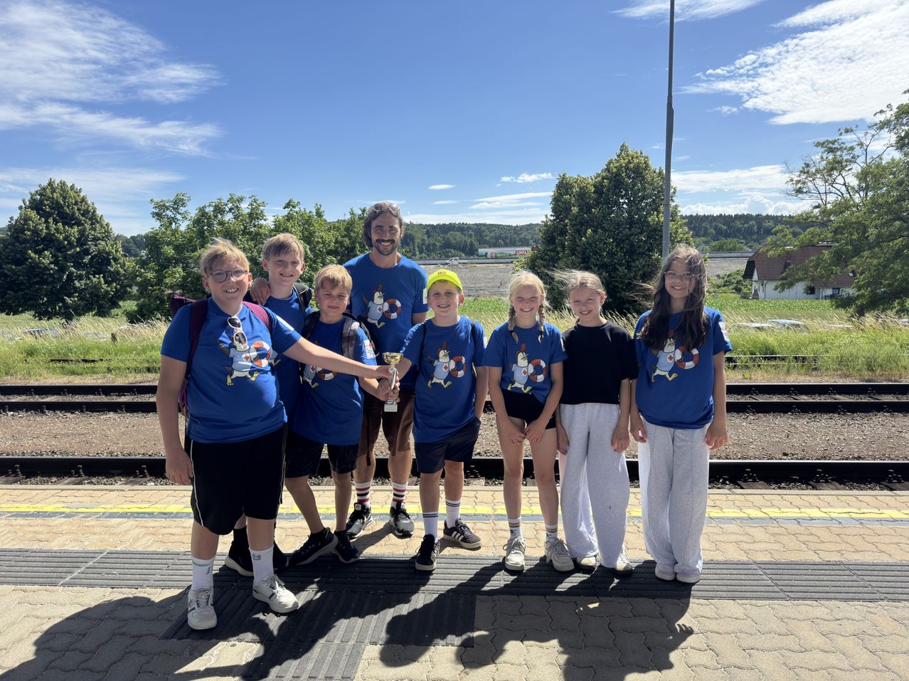 Eine Gruppe von Kindern und einem Mann steht auf einer Plattform in übereinstimmenden blauen T-Shirts. Sie posieren für ein Foto und halten eine Trophäe. Dahinter befindet sich eine Bahnlinie und einige Bäume.