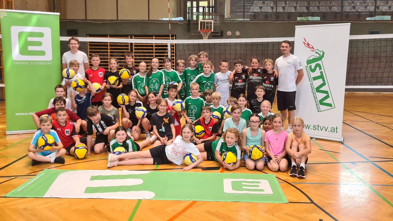 Eine Gruppe junger Athleten in grünen und weißen Uniformen posiert mit Volleyballs in einer Sporthalle. Ein Mann in einem weißen Shirt steht hinter dem Netz.