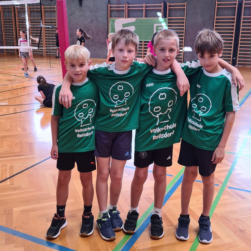 Vier Jungen in grünen T-Shirts mit 'Volksschule Rollsdorf' stehen in einem Gymnastikraum. Sie lächeln und einer hat seinen Arm um den anderen gelegt. Hinter ihnen ist ein Volleyballnetz und eine Person zu sehen, die auf dem Boden sitzt.