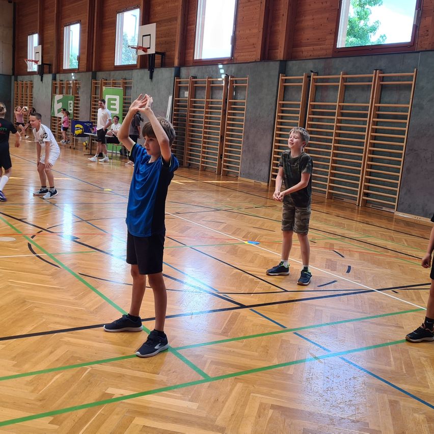 Eine Gruppe von Kindern spielt Basketball auf einem Indoor-Spielfeld. Ein Junge springt mit erhobenen Händen, während ein anderer hinter ihm steht.