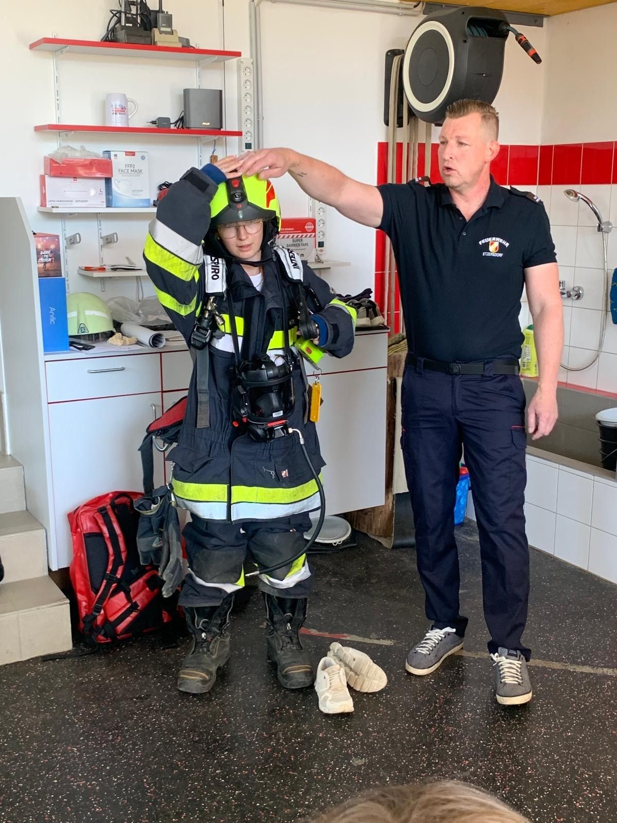 Ein Feuerwehrmann in Uniform steht mit einem Feuerwehrchef in einer Feuerwache, mit Ausrüstung, Gerät und einem roten Rucksack in der Nähe.