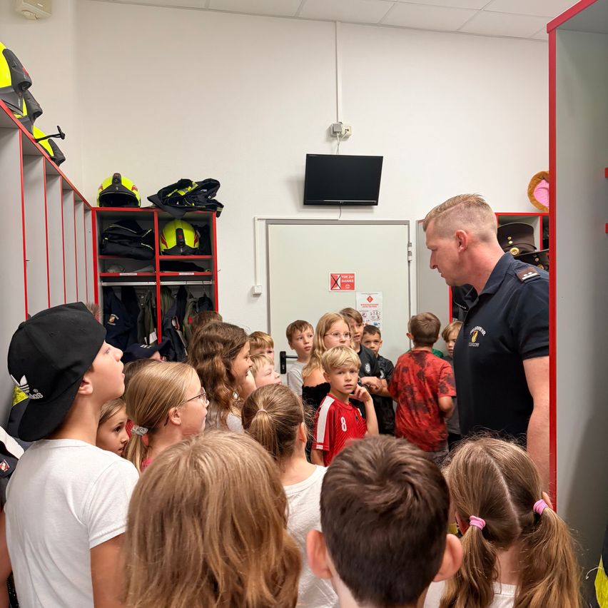 Ein Feuerwehrmann spricht mit einer Gruppe von Kindern in einer Feuerwache, mit Schutzausrüstung auf den Regalen im Hintergrund.