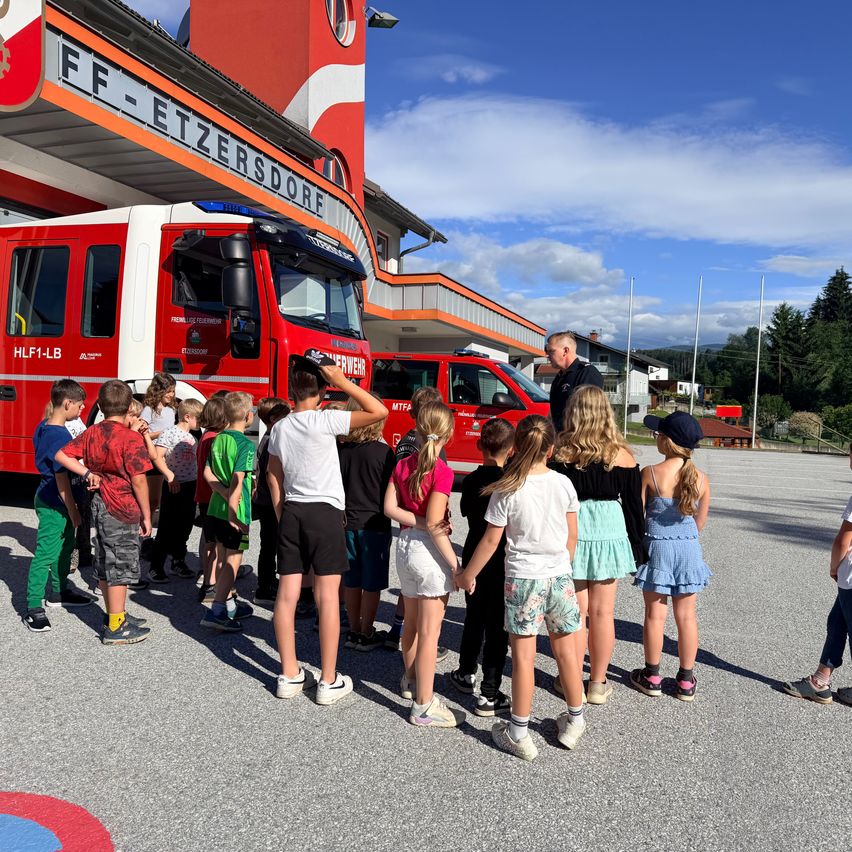 Eine Gruppe von Kindern und Erwachsenen steht vor einem Feuerwehrauto bei einer Feuerwache, wobei ein Mann ihnen etwas erklärt.