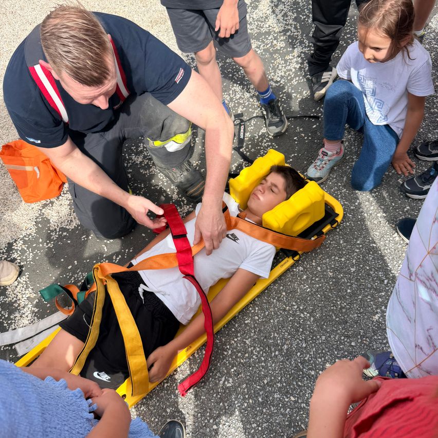 Eine Person liegt auf einer Trage, während ein Erwachsener in einer blauen Uniform sie mit einem Gurtzeug sichert. Mehrere Kinder beobachten in der Nähe.