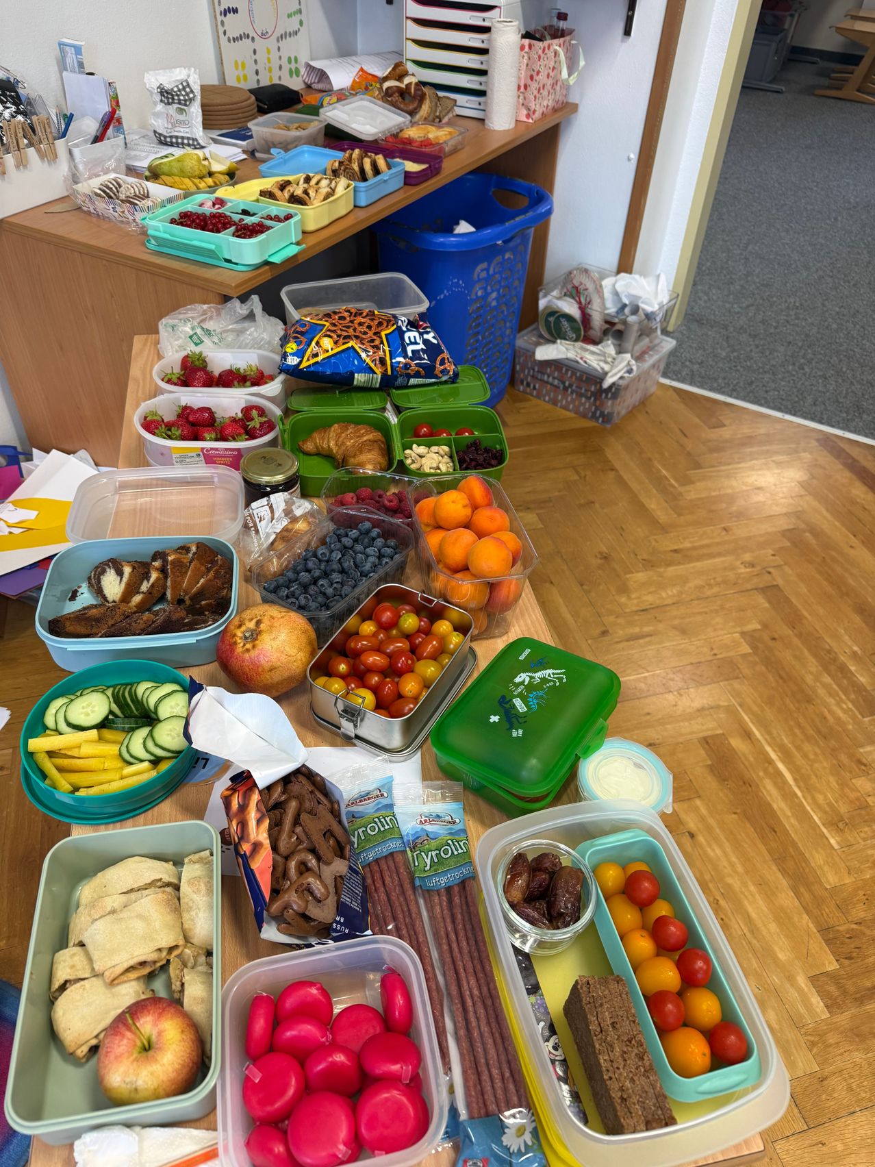 A table filled with various food items in plastic containers, including fruits, vegetables, and snacks. There are also some bread rolls and a bag of pretzels.