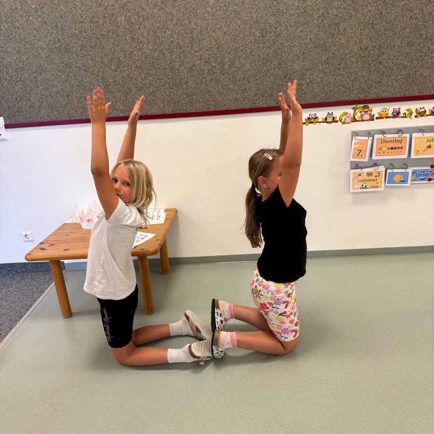 Two young girls are doing yoga poses in a classroom with a wooden table and wall decorations.