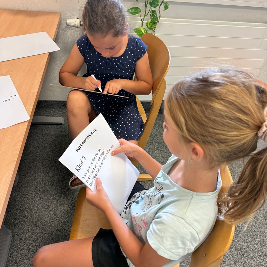 Two young girls are sitting on chairs in a classroom, one is holding a paper with the text 'Kind 2 Partnerdikat' while the other is writing on a piece of paper.