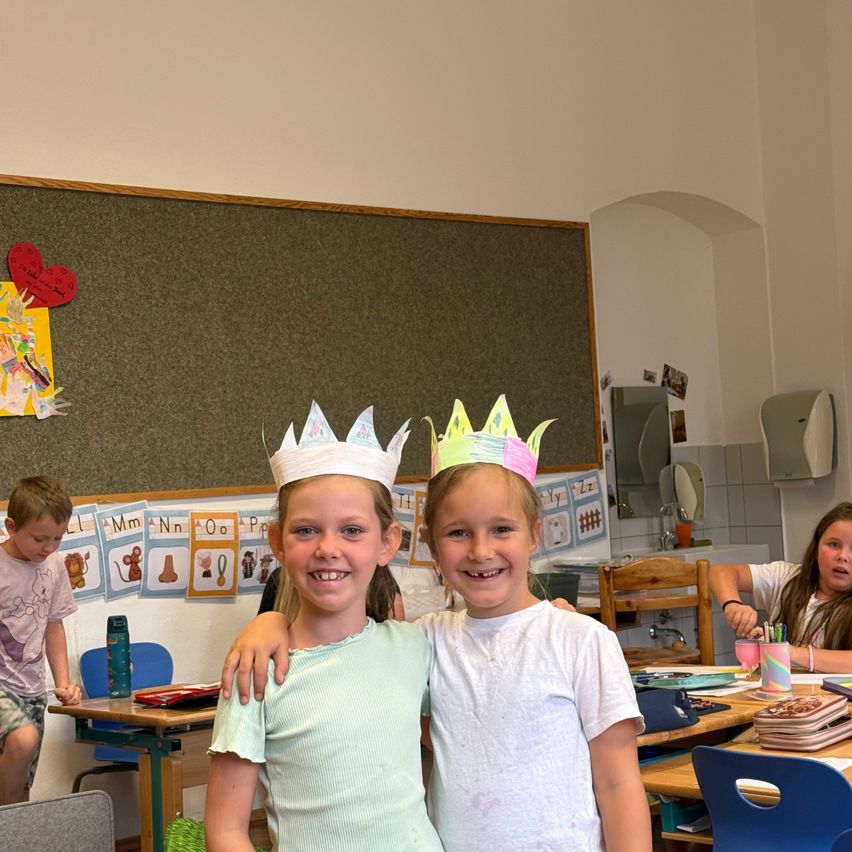Two young girls wearing paper crowns smile for a photo in a classroom with a bulletin board in the background.