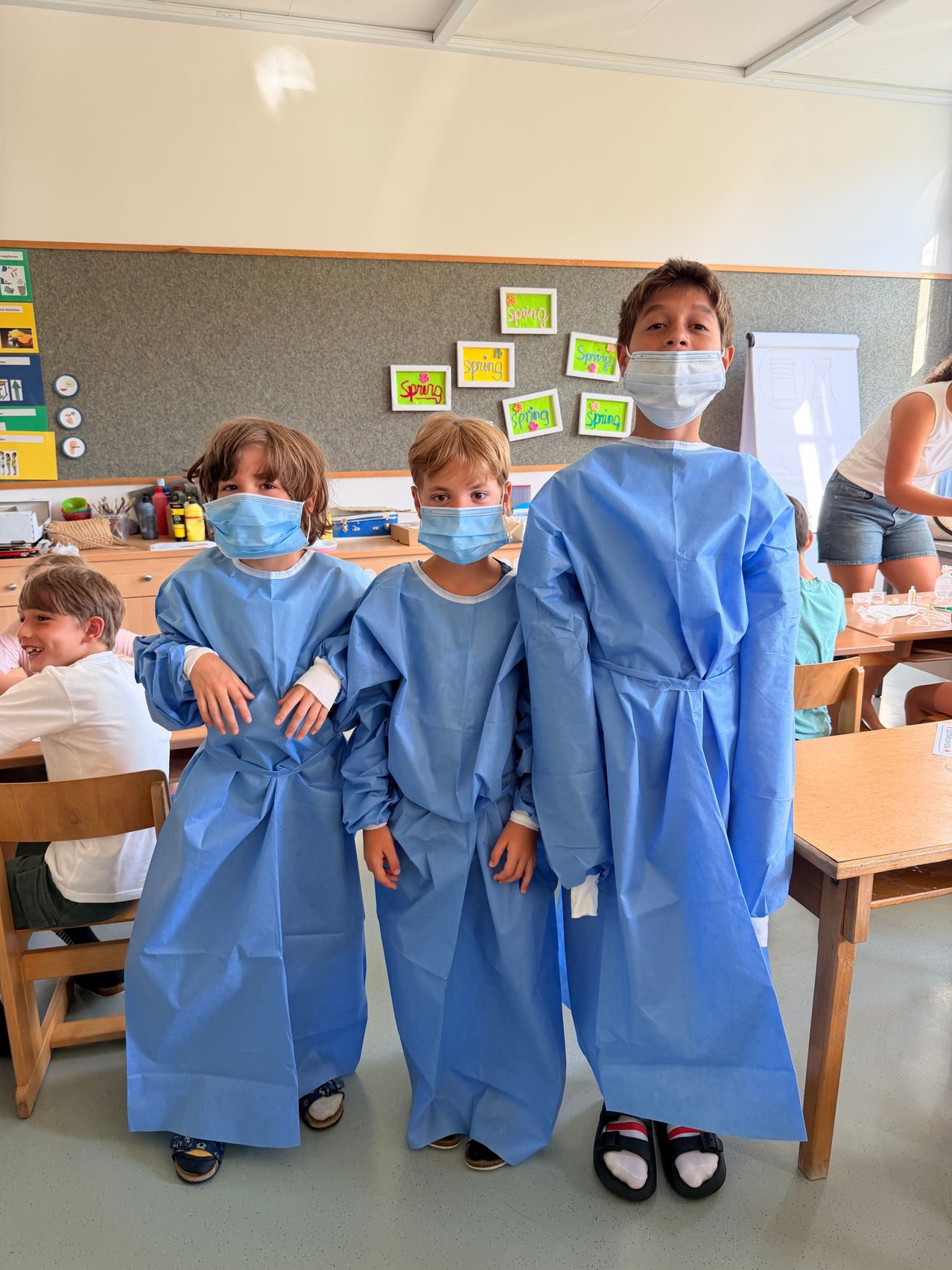 Three children in blue medical gowns and masks stand together in a classroom, with other students and a teacher in the background.