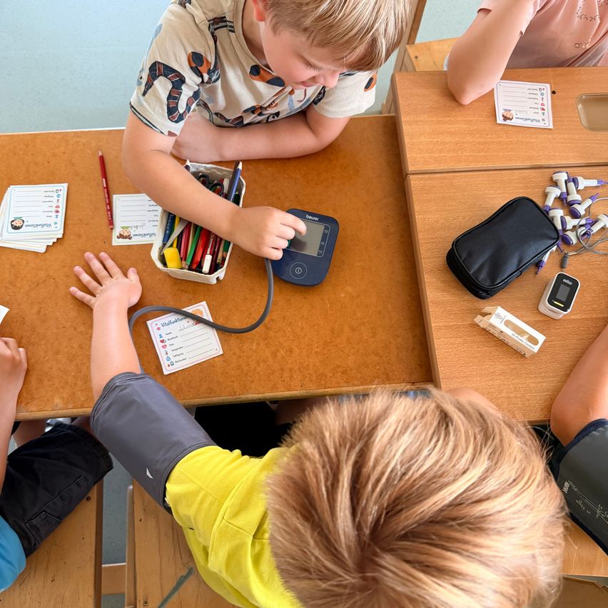 A young boy is measuring his blood pressure using a cuff and a monitor while sitting at a desk with other children.