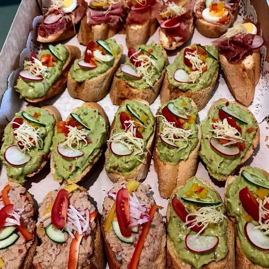 Eine Schachtel gefüllt mit verschiedenen Arten von Bruschetta, einschließlich Avocado-, Tomaten- und Käsebelägen, in einer ordentlichen Reihe angeordnet.
