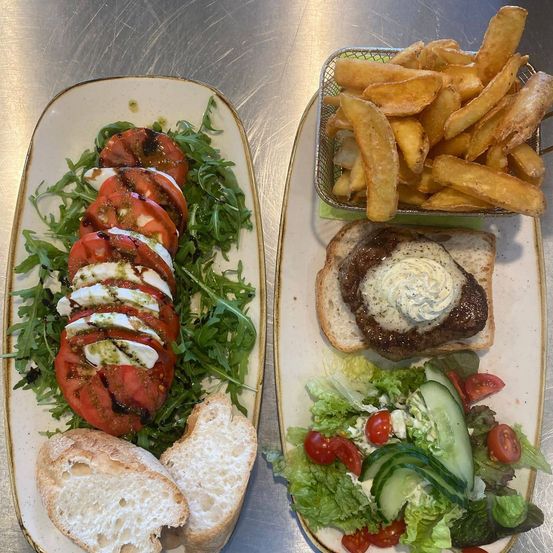 Zwei Teller mit Essen: einer mit einem Caprese-Salat aus Tomaten, Mozzarella und Basilikum und ein anderer mit einem Burger, Pommes und einem Salat mit Tomaten und Gurken.