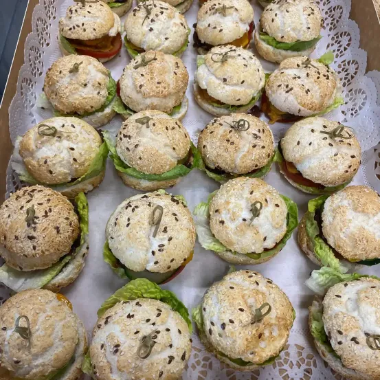 Ein Tablett gefüllt mit kleinen Sesambrötchen, jedes belegt mit Salat, Tomate und einem kleinen Essiggurken. Die Brötchen sind ordentlich auf einem weißen Spitzentuch angeordnet.