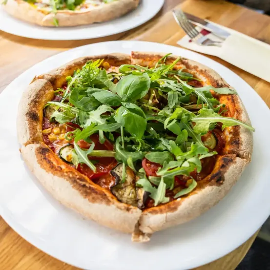 Eine frisch gebackene Pizza mit Rucola und Gemüse auf einem weißen Teller auf einem Holztisch.