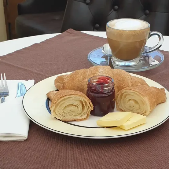 Ein Teller mit drei Croissants, Butter und einer Glasdose mit Marmelade steht auf einem Tisch, begleitet von einer Tasse Kaffee mit Schaum auf einer Untertasse.