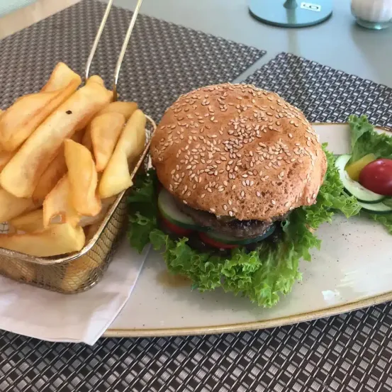 Ein Burger mit Sesamkörnern auf einem Brötchen, begleitet von einer Portion Pommes und einem Salat mit Salat, Tomate und Gurke.