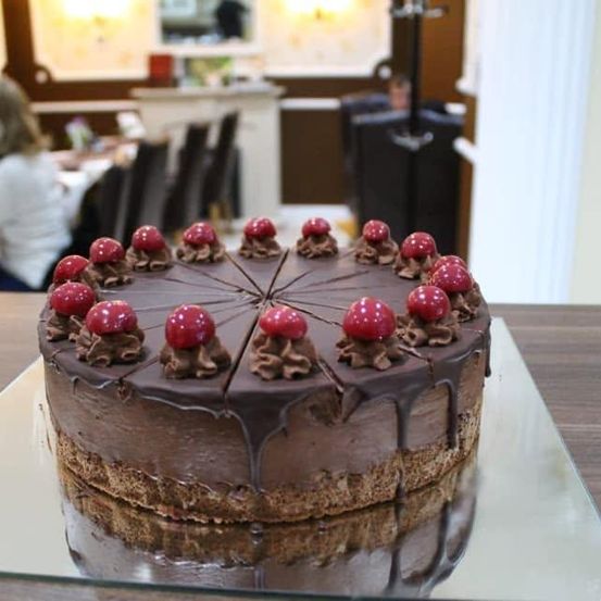 Ein Schokoladenkuchen mit roten Kirschen auf einem Glasständer in einem Restaurant. Eine Frau sitzt im Hintergrund an einem Tisch.