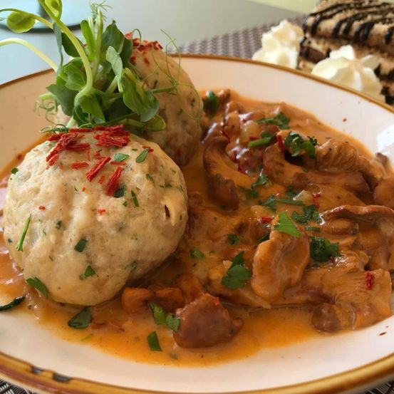 Ein Teller mit Pilzgericht in cremiger Sauce, garniert mit Petersilie und roten Chiliflocken, begleitet von zwei Knödeln und einer Seite Schlagsahne.