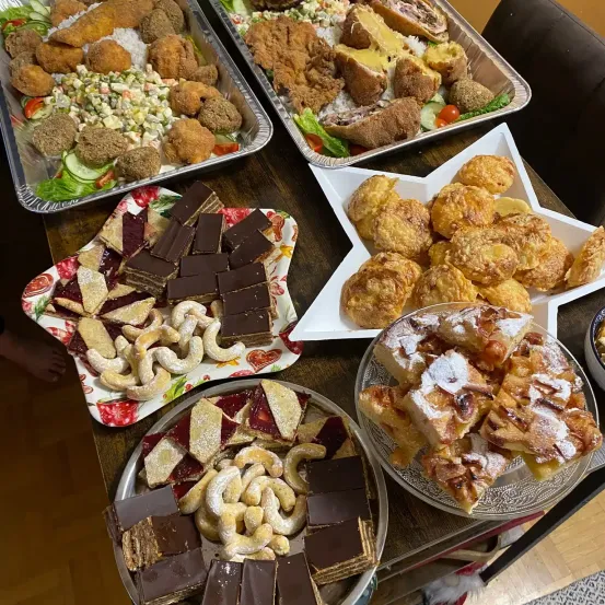 Ein Tisch mit verschiedenen Desserts wie Schokoladenquadraten, Gebäck und Obsttörtchen. Es sind auch Schalen mit frittiertem Essen und Salaten vorhanden.