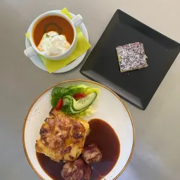 Eine Mahlzeit mit einer Tasse Suppe, einem Teller mit Gemüse, einem Dessert und einem Teller mit Fleisch mit Soße.