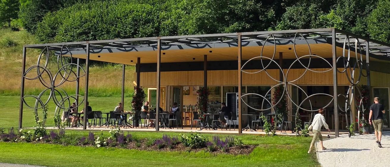Ein modernes Gebäude mit kreisförmigen Designs steht in einem Park, wo Menschen spazieren und auf Stühlen unter einem Baldachin sitzen. Im Hintergrund befindet sich ein Schloss auf einem Hügel mit üppiger Vegetation.