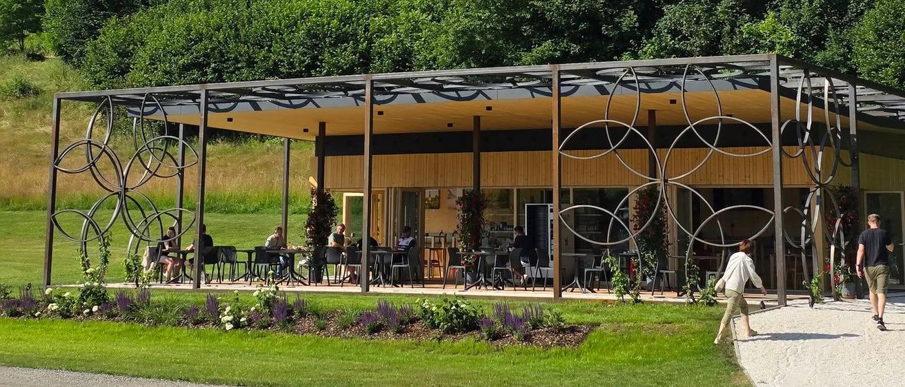 Ein modernes Gebäude mit kreisförmigen Designs steht in einem Park, wo Menschen spazieren und auf Stühlen unter einem Baldachin sitzen. Im Hintergrund befindet sich ein Schloss auf einem Hügel mit üppiger Vegetation.
