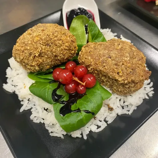Ein schwarzer Teller mit zwei frittierten Kroketten, roten Beeren und einem Blatt mit schwarzer Sauce auf einem Bett aus Reis.