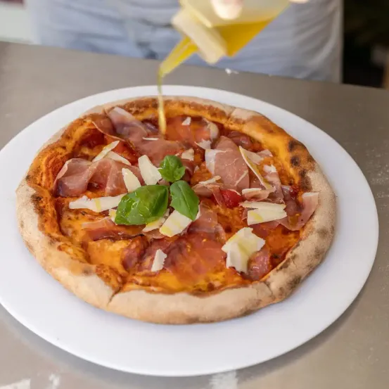 Eine Pizza mit Belag aus Schinken, Käse und Basilikum wird auf einem weißen Teller mit Olivenöl beträufelt.