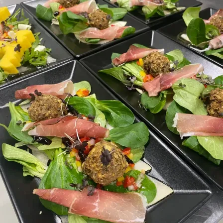 Eine Nahaufnahme von mehreren schwarzen Tellern mit einem Salatgericht. Der Salat besteht aus grünen Blättern, Fleisch und Gemüse. Das Gericht ist mit schwarzen Samen garniert.
