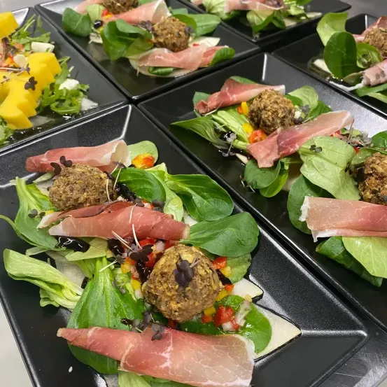 Eine Nahaufnahme von mehreren schwarzen Tellern mit einem Salatgericht. Der Salat besteht aus grünen Blättern, Fleisch und Gemüse. Das Gericht ist mit schwarzen Samen garniert.