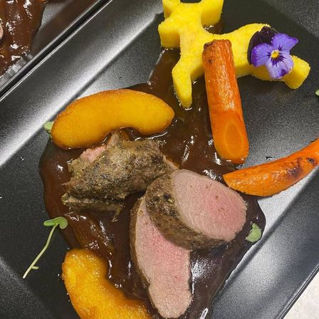 Ein Gourmetgericht auf einem schwarzen Teller besteht aus Fleischscheiben mit einer reichhaltigen braunen Sauce, begleitet von gerösteten Karotten, gelbem Kürbis und einer violetten Blume als Garnierung.