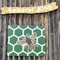 Ein hölzernes Schild mit einem Bienenwabenmuster und einer Biene-Illustration ist an einer Holzwand befestigt. Darunter steht ein gelbes Schild mit dem Wort 'Rauch' in weiß.