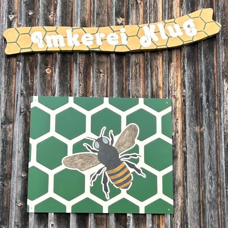 Ein hölzernes Schild mit einem Bienenwabenmuster und einer Biene-Illustration ist an einer Holzwand befestigt. Darunter steht ein gelbes Schild mit dem Wort 'Rauch' in weiß.