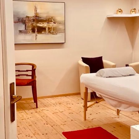 Ein Raum mit Holzboden, einer Wand mit einem Gemälde, einem Stuhl und einem Massagetisch mit Kissen. Ein Regal hält zwei goldene Objekte.