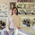 Eine Frau steht in einem Laden mit einer weißen Theke und lächelt. Hinter ihr befinden sich Regale mit Produkten und ein Schild an der Wand. Links stehen Flaschen mit Wein.