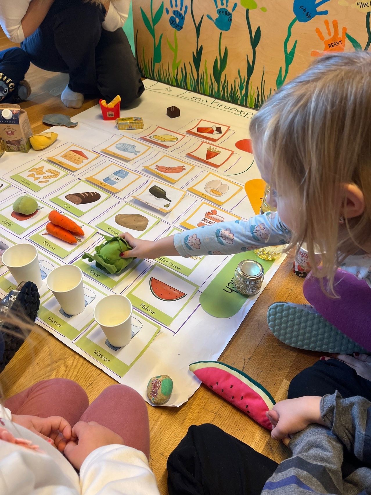 Ein kleines Mädchen interagiert mit einem farbenfrohen Lernspiel auf einem Holzboden, das Lebensmittel und Getränke zeigt, während ein anderes Kind in der Nähe zusieht.
