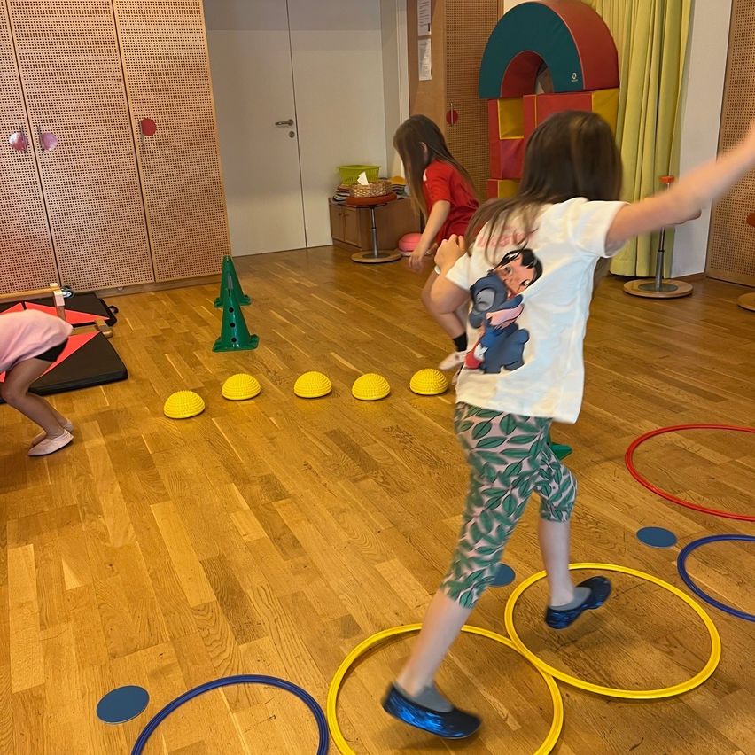 Mehrere Kinder spielen auf einem Holzboden mit gelben und blauen Ringen, Kegeln und Bällen. Ein Mädchen in einem weißen T-Shirt springt über einen gelben Ring. Hinter ihr geht ein Mädchen in einem roten T-Shirt mit einem Ball.