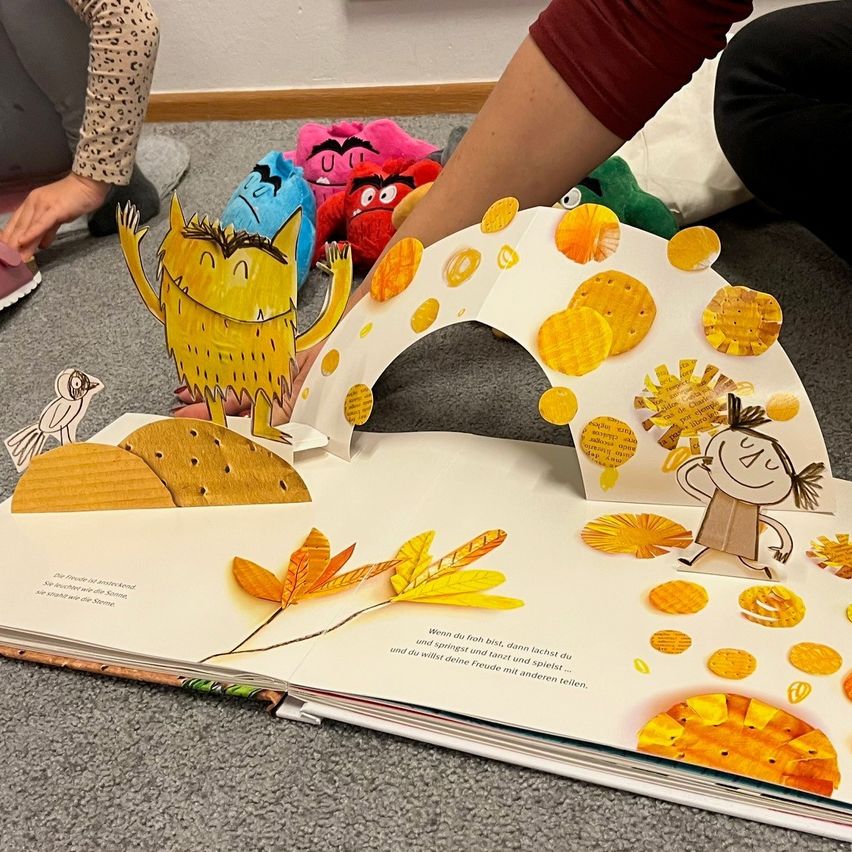 Ein offenes Kinderbuch mit einer Papierausschnittszene, die einen Monster, ein Mädchen und einen Tunnel aus goldenen Blättern zeigt. Das Buch liegt auf einem Teppichboden mit verstreuten Spielzeugen.