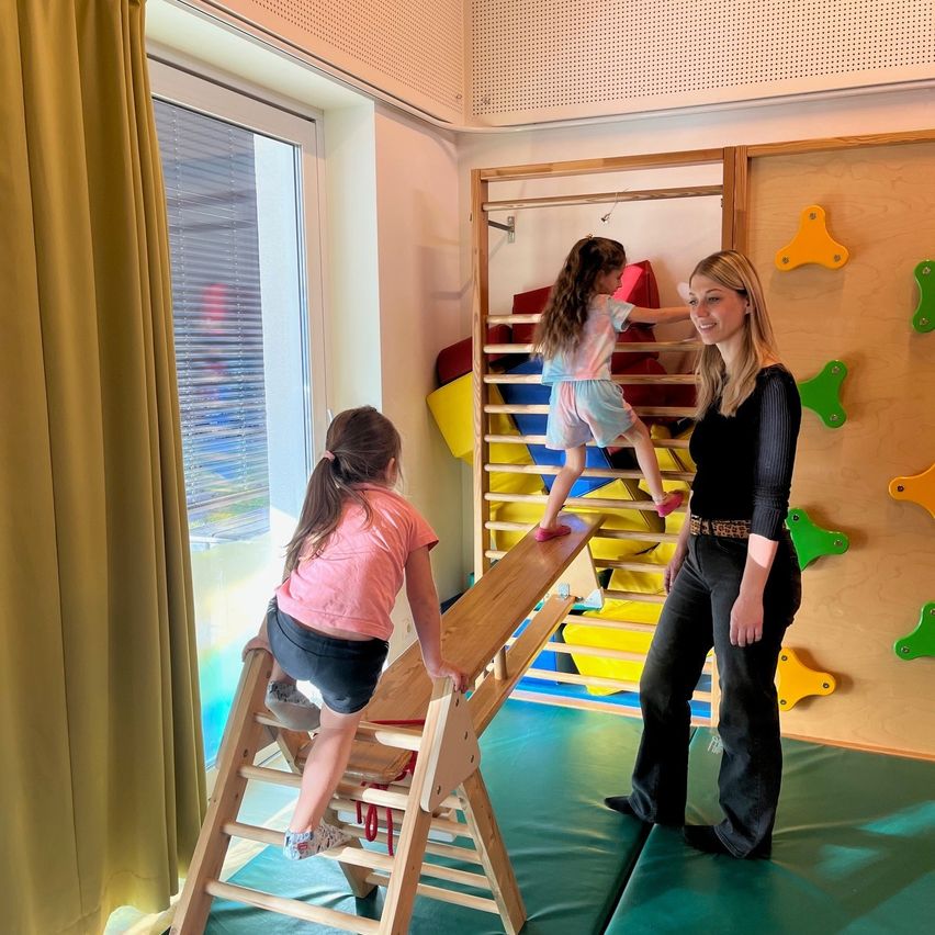 Eine Frau und zwei Mädchen spielen auf einem Wippgerät und Klettergerüst in einem Spielzimmer. Die Frau lächelt und beobachtet die Mädchen.