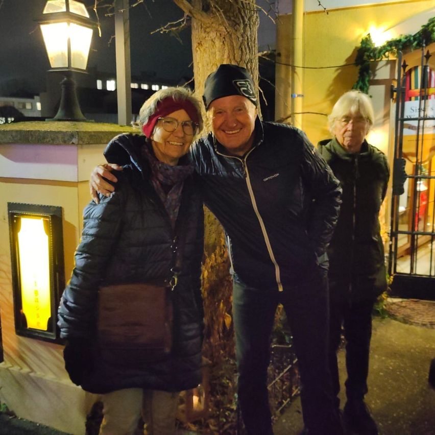 Drei Menschen stehen nachts draußen und lächeln. Ein Mann in der Mitte hat seinen Arm um eine Frau gelegt, und eine weitere Frau steht in der Nähe. Sie stehen neben einem Baum und einer Laterne.