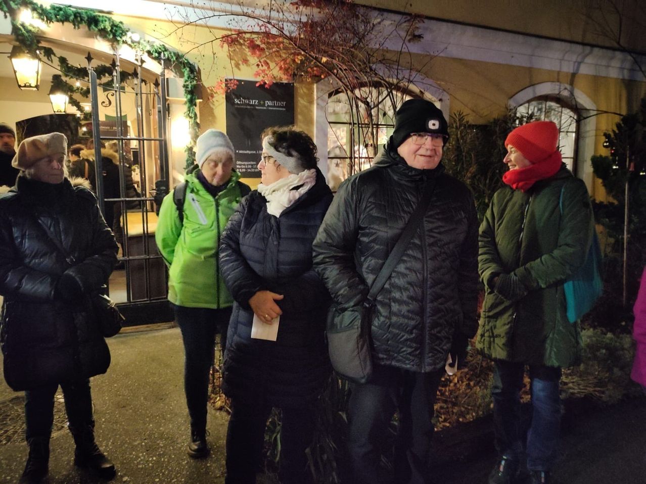 Vier Menschen, alle warm gekleidet, stehen nachts vor einem Gebäude. Sie lächeln und scheinen sich zu unterhalten. Ein Mann trägt einen schwarzen Hut und Brille. Hinter ihnen steht ein Baum mit roten Blättern und ein Schild.