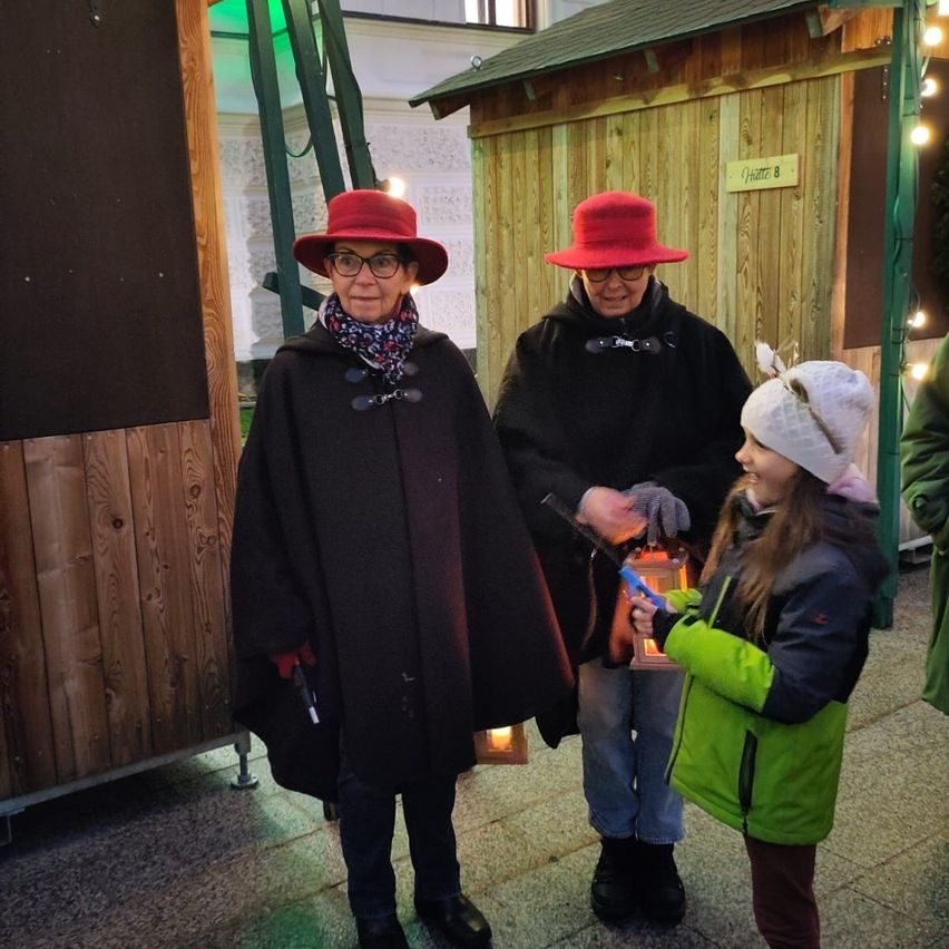 Drei Menschen stehen vor einer Holzhütte. Zwei Erwachsene tragen Hüte und Mäntel, während ein junges Mädchen eine Laterne hält.