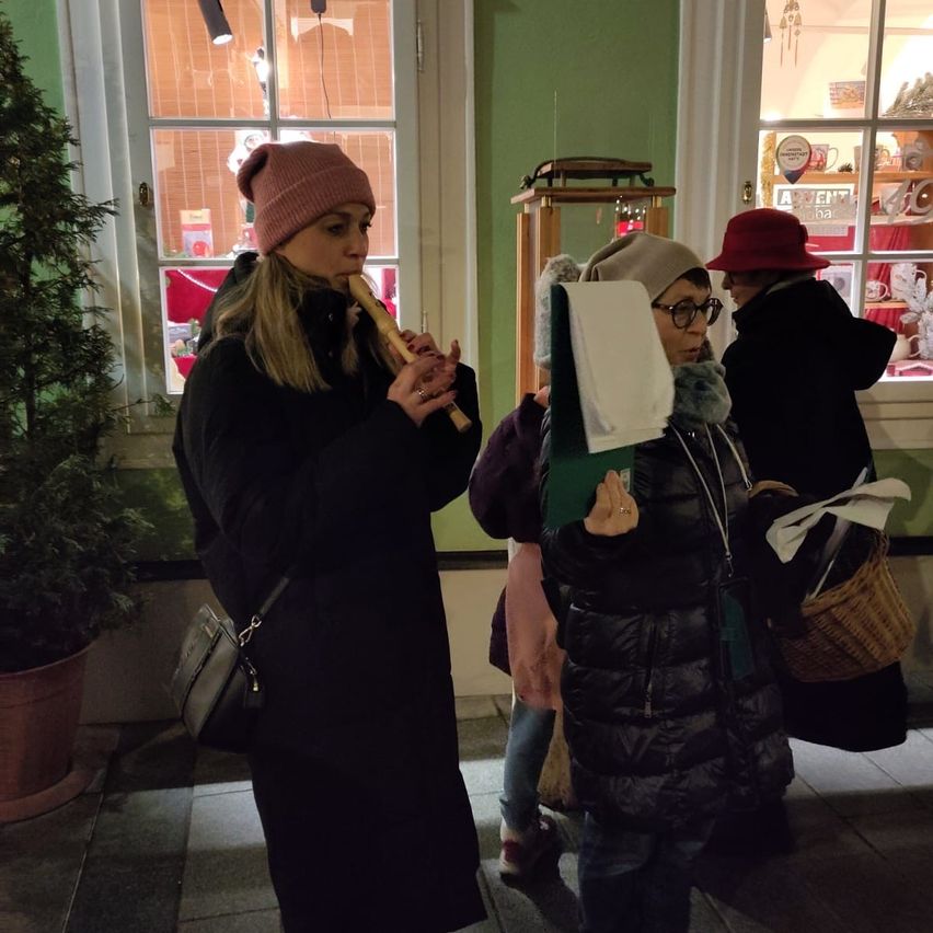 Eine Frau mit einer rosa Mütze spielt eine Flöte, während andere stehen, eine hält einen Korb, in einem Raum mit grünen Wänden und einem Topf.