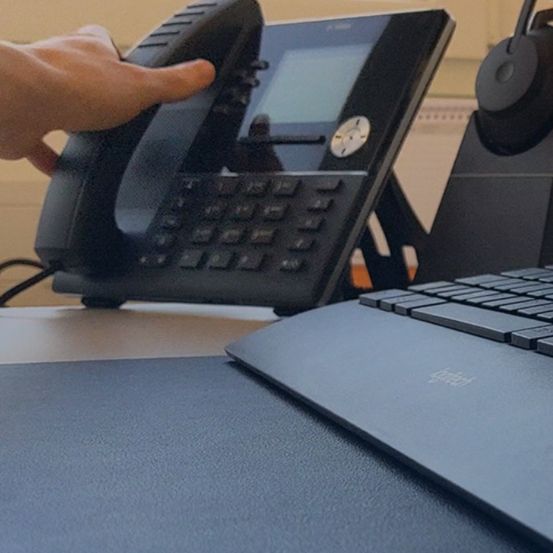 Eine Hand hält ein schwarzes Telefon mit einem digitalen Bildschirm und Tasten auf einem Schreibtisch, begleitet von einer Tastatur.